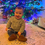 Austin joined the competition — help win amazing prizes! baby, child, smiling, sitting, sweater, jeans, boots, christmas_tree, ornaments, lights, presents, rug, indoor, holiday, festive, decorations, happy, cute, home, floor