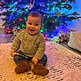 baby, child, smiling, sitting, sweater, jeans, boots, christmas_tree, ornaments, lights, presents, rug, indoor, holiday, festive, decorations, happy, cute, home, floor