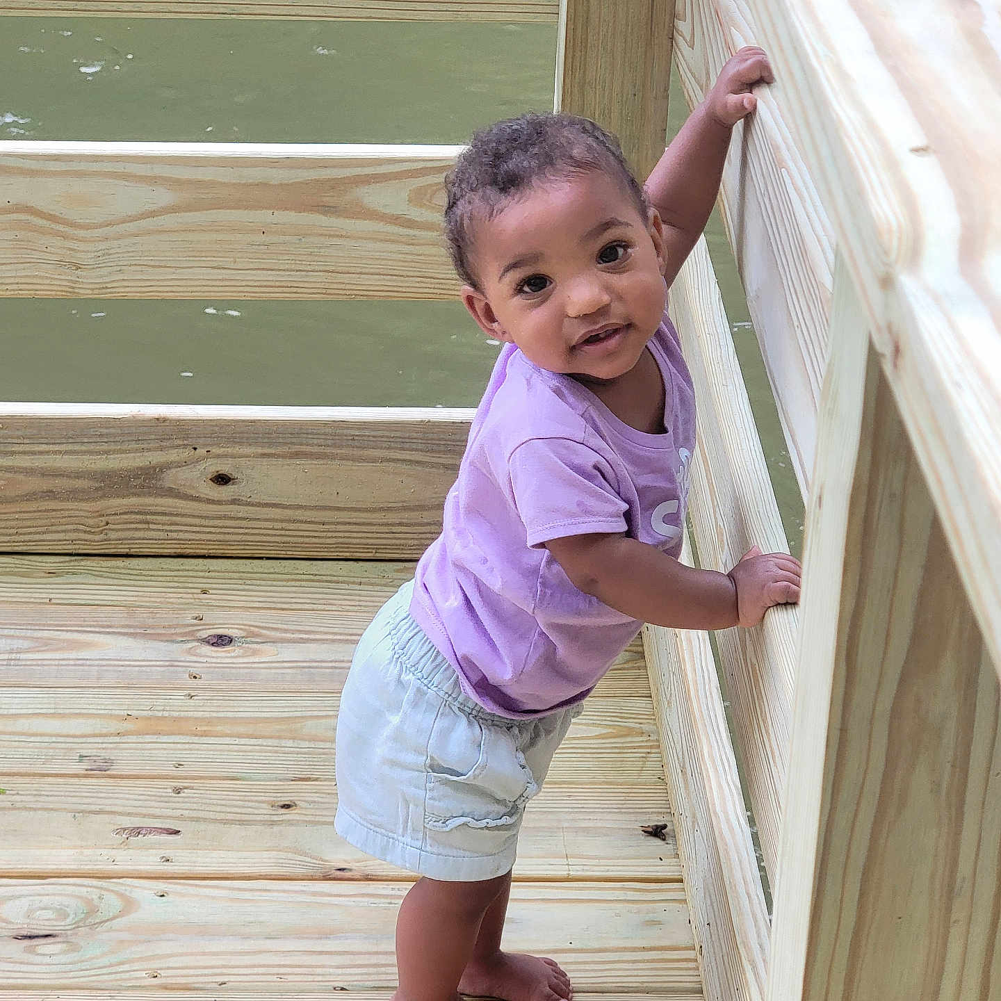 Kei'Lani joined the competition — help win amazing prizes! baby, barefoot, child, curious, cute, daylight, light_shorts, looking_up, nature, outdoor, person, portrait, purple_shirt, railing, smiling, standing, toddler, water, wood, wooden_deck