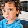 child, curly_hair, blue_eyes, sweatshirt, indoor, portrait, young_child, face, person, casual_clothing, modern_interior, fireplace, thoughtful, cute, kid, seated, expression, closeup, natural_light, hairstyle