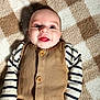 baby, smiling, face, infant, striped_shirt, vest, buttoned_clothing, blanket, checkered_pattern, cozy, happy, lying_down, child, cute, portrait, indoors, person, warm_colors, soft_texture, expressive