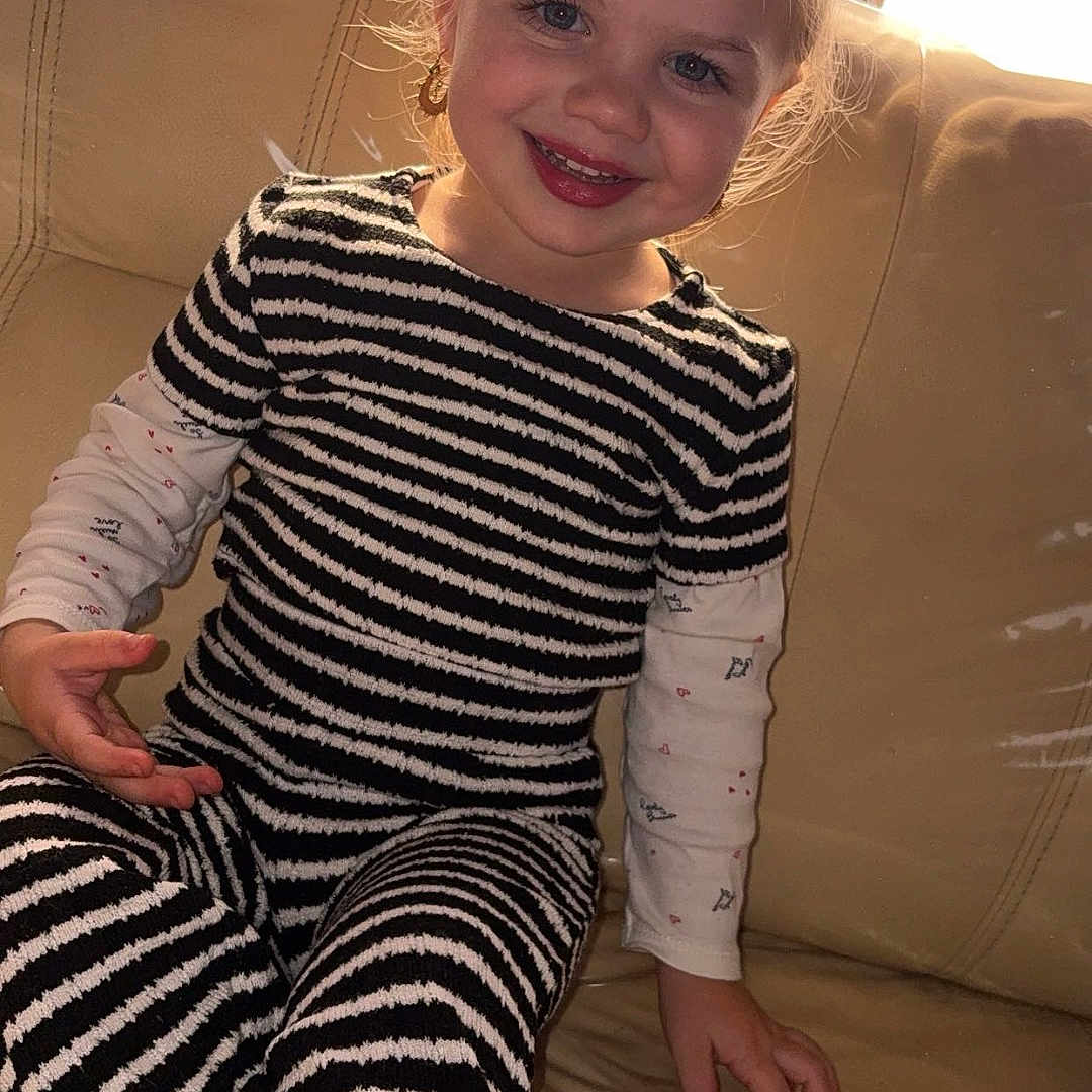 Carla a rejoint le concours — aidez-le/la à gagner de superbes lots ! blonde_hair, blue_eyes, casual_clothing, child, couch, cozy, cute, earrings, furniture, happy, home, indoor, long_sleeve_shirt, natural_light, portrait, sitting, smiling, striped_clothing, toddler, young_child