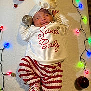 Anzal is registered to the contest to win money with this photo: baby, sleeping, christmas, holiday, festive, lights, ornaments, red, white, striped_pants, sweater, hat, smiling, cute, infant, cozy, indoors, celebration, decoration, lying_down