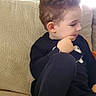 child, boy, toddler, portrait, sitting, couch, indoors, home, family_photos, curly_hair, profile, thoughtful, casual_clothing, black_outfit, shirt, pants, barefoot, soft_lighting, living_room, cozy