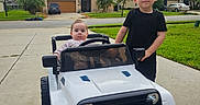 Yusuf Eren is registered to the contest to win money with this photo: children, baby, toddler, toy_car, jeep, ride_on_vehicle, outdoor, driveway, suburb, house, lawn, palm_trees, portrait, siblings, sitting, standing, playtime, smiling, vehicle, daylight