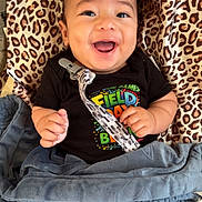 Zayan is registered to the contest to win money with this photo: baby, smiling, infant, blanket, leopard_print, clothing, pacifier_clip, happy, face, person, indoors, cute, child, lying_down, black_shirt, blue_blanket, patterned_cushion, cheerful, small_hands, bright_eyes