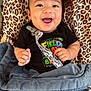 baby, smiling, infant, blanket, leopard_print, clothing, pacifier_clip, happy, face, person, indoors, cute, child, lying_down, black_shirt, blue_blanket, patterned_cushion, cheerful, small_hands, bright_eyes