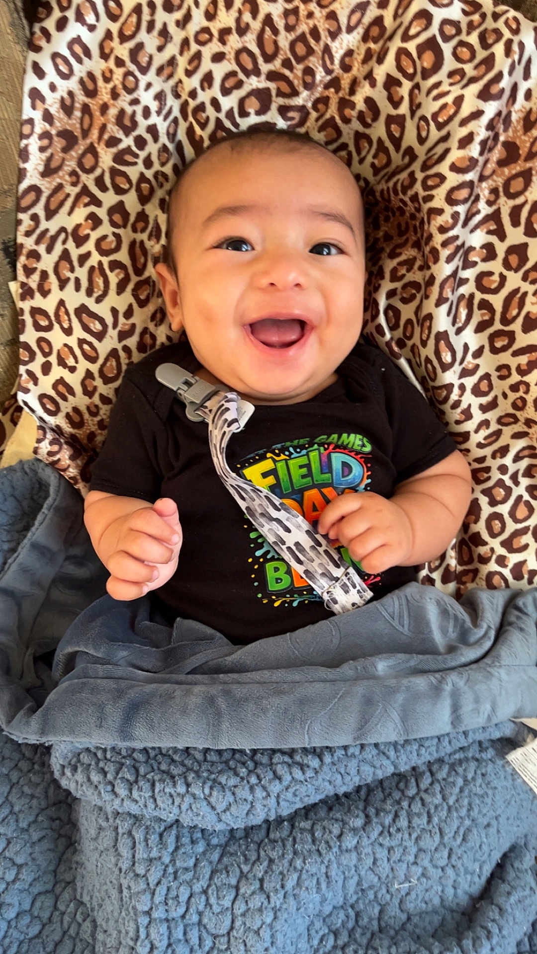 Zayan is registered to the contest to win money with this photo: baby, smiling, infant, blanket, leopard_print, clothing, pacifier_clip, happy, face, person, indoors, cute, child, lying_down, black_shirt, blue_blanket, patterned_cushion, cheerful, small_hands, bright_eyes