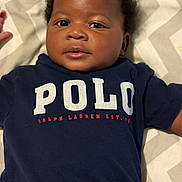 Denim is registered to the contest to win money with this photo: baby, child, face, person, infant, lying_down, blue_shirt, polo, curly_hair, hand, blanket, pattern, cute, portrait, skin, young, clothing, indoors, expression, toddler