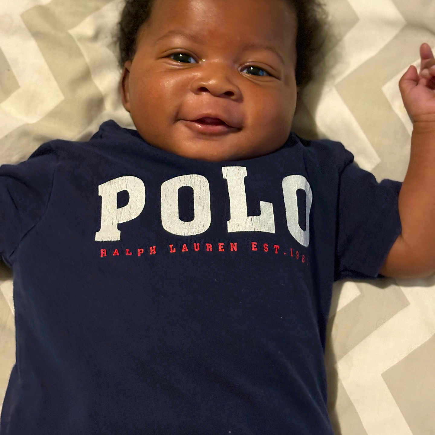 Denim is registered to the contest to win money with this photo: baby, smiling, infant, face, clothing, polo_shirt, navy_blue, lying_down, blanket, pattern, cute, happy, child, portrait, one_person, indoor, skin, head, hair, toddler