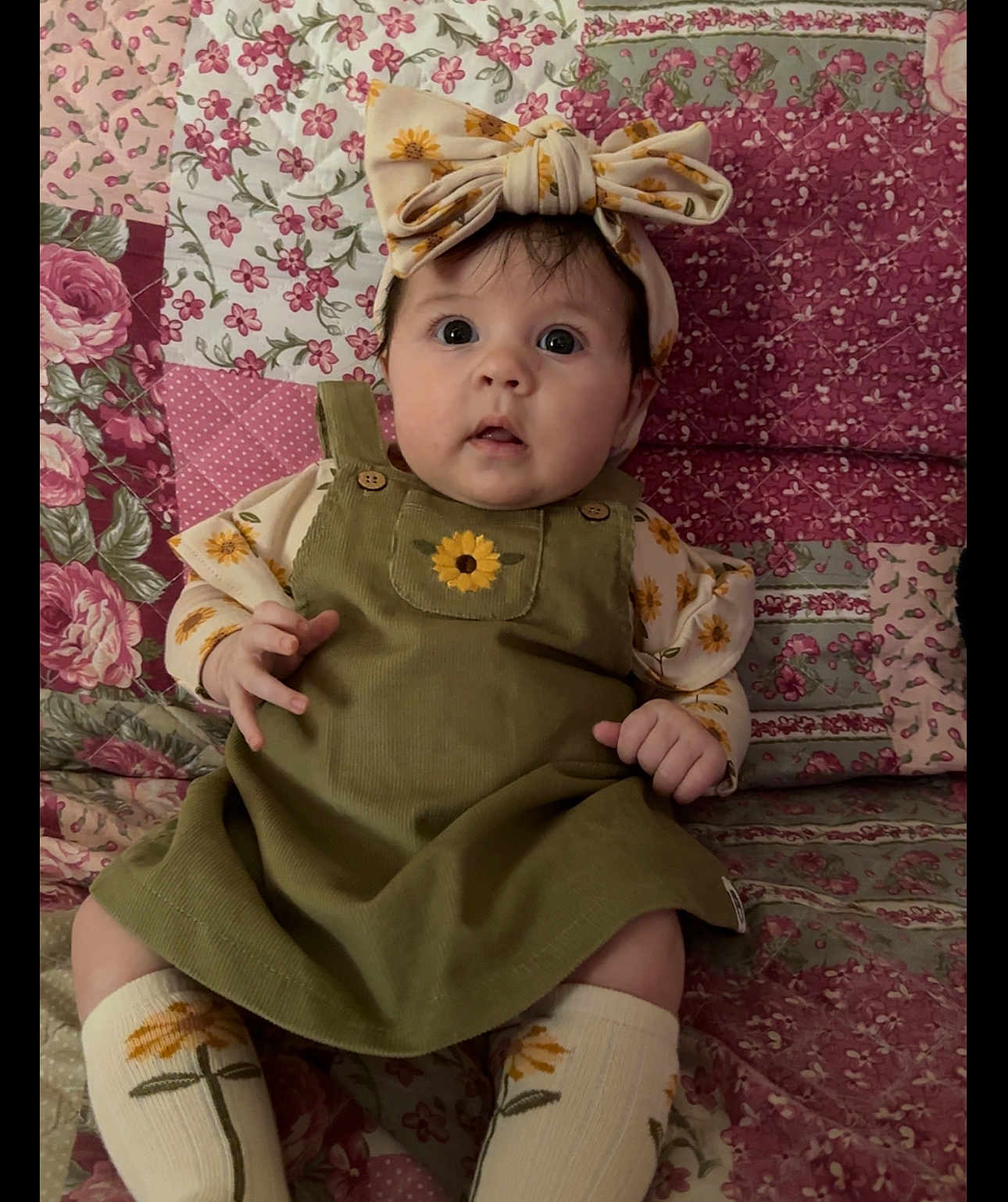Rosemary is registered to the contest to win money with this photo: baby, child, infant, face, headband, flower, sunflower, dress, green_dress, patterned_socks, floral_socks, floral_background, quilt, cute, portrait, indoors, seated, hands, clothing, expression