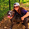 woman, dog, outdoor, smiling, sunglasses, cap, fence, gravel, collar, happy, pet, blue_eyes, sunlight, casual_clothing, nature, portrait, friendship, animal, human, leisure