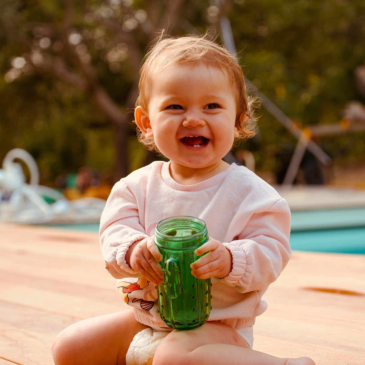 Jeanne participe au concours pour gagner de l'argent avec cette photo : toddler, child, smiling, sitting, green_cup, wooden_deck, outdoor, daylight, happy, short_hair, casual_clothing, diaper, nature, trees, pool, baby, cute, playful, person, portrait