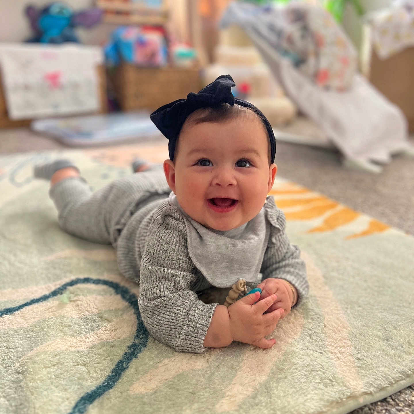 Olivia is registered to the contest to win money with this photo: baby, carpet, child, cozy, cute, expression, floor, gray_clothing, hands, happy, headband, home, indoor, infant, person, playful, rug, smiling, soft_lighting, toy