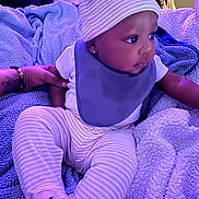 Baby Eli is registered to the contest to win money with this photo: baby, bed, bib, blanket, bracelet, closet, cozy, cute, earring, hand, hat, indoor, infant, onesie, plush_texture, portrait, purple_lighting, striped_pants, tattoo, toddler