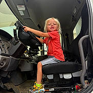 Sebastian is registered to the contest to win money with this photo: child, blonde_hair, smiling, sitting, driver_seat, steering_wheel, vehicle_interior, orange_shirt, sneakers, yellow_socks, happy, dashboard, carpet, footwear, seat, window, door, hand, floor, toy_pattern