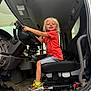 child, blonde_hair, smiling, sitting, driver_seat, steering_wheel, vehicle_interior, orange_shirt, sneakers, yellow_socks, happy, dashboard, carpet, footwear, seat, window, door, hand, floor, toy_pattern