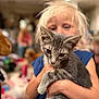 Sebastian is registered to the contest to win money with this photo: child, kitten, cat, holding, indoor, blond_hair, gray_cat, pet, cute, young_child, person, animal, portrait, furniture, toy, blurred_background, living_room, casual_clothing, happy, domestic