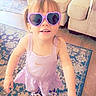 toddler, child, girl, sunglasses, heart_shaped, dress, purple, shoes, indoor, carpet, rug, smiling, happy, furniture, couch, storage_bin, flooring, hair, person, home