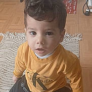 Kassim a rejoint le concours — aidez-le/la à gagner de superbes lots ! big_eyes, casual_clothing, child, christmas_tree, curly_hair, cute, home_interior, indoor, kid, kneeling, looking_up, mustard_sweater, ornament, portrait, puma_logo, rug, sitting, toddler, wooden_floor, young_child