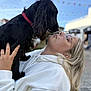 affection, black_dog, blonde_hair, candid, close_up, daylight, dog, face, flags, friendly, glasses, holding, love, nose_to_nose, outdoor, person, pet, portrait, sky, white_hoodie