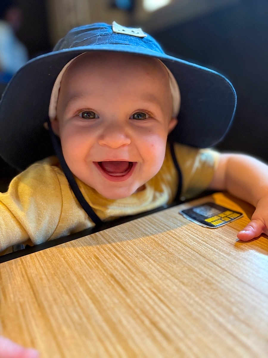 Malachi is registered to the contest to win money with this photo: baby, child, smiling, blue_hat, yellow_shirt, table, happy, face, portrait, indoor, young_child, cute, person, closeup, headwear, infant, baby_hands, bright_eyes, cheerful, wooden_surface