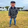 child, boy, grass, field, jacket, jeans, boots, cap, smiling, outdoor, caravan, van, vehicle, nature, cloudy_sky, standing, portrait, casual_clothing, happy, daytime