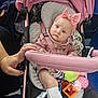 baby, stroller, pink, headband, bow, toy, hand, socks, child, seatbelt, comfort, indoor, person, cute, infant, expression, clothing, sitting, blue_eyes, soft_toys