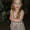 blonde_hair, blue_eyes, books, bookshelf, child, crossed_arms, dress, flash_photography, floral_dress, girl, home, indoor, living_room, long_hair, pink_clothing, portrait, serious_expression, shoes, standing, toys