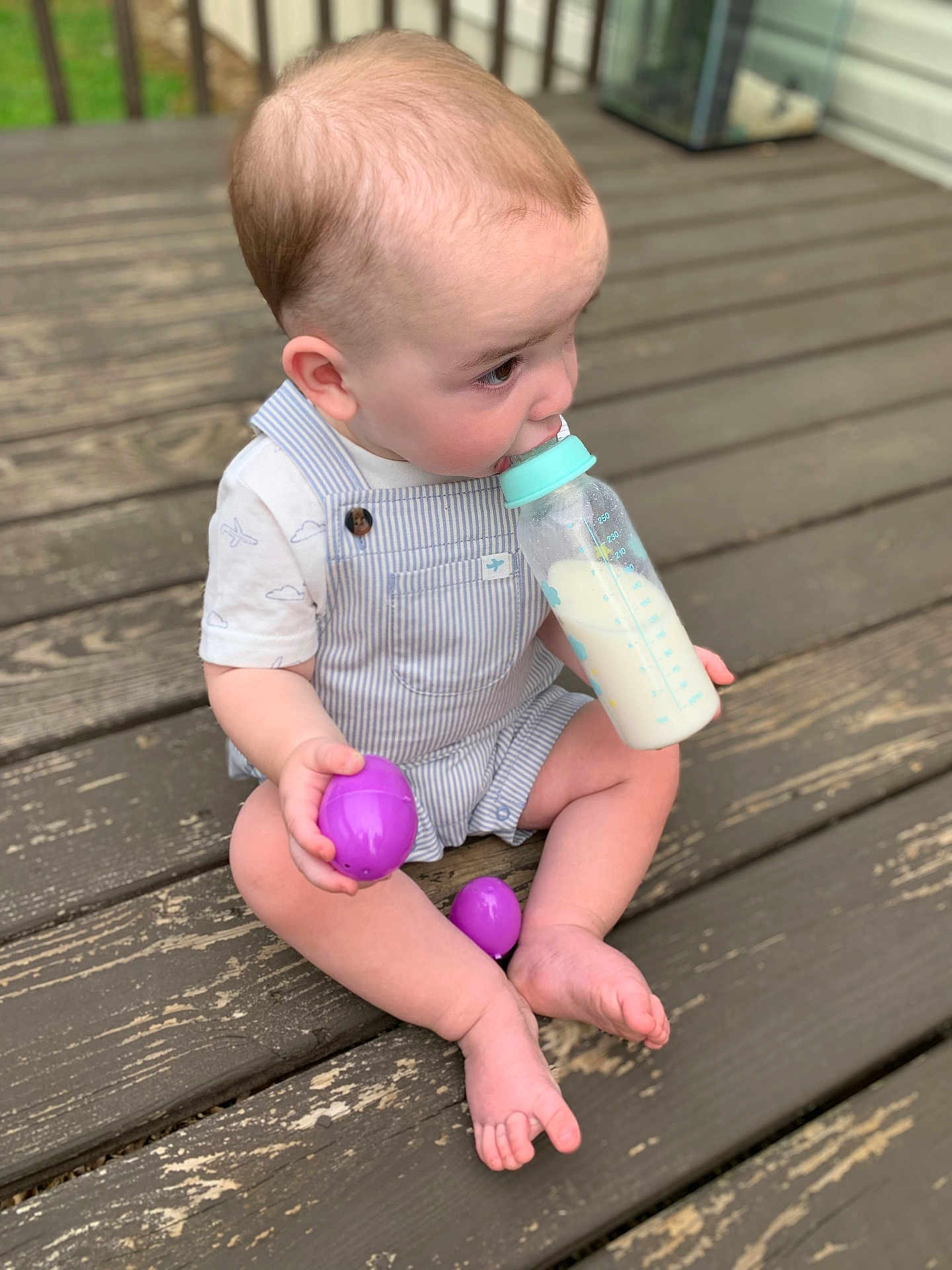 Colton is registered to the contest to win money with this photo: baby, child, milk_bottle, purple_eggs, overalls, striped_clothing, wooden_deck, outdoor, sitting, barefoot, cute, infant, playful, side_view, summer, daylight, young_child, holding_object, casual, relaxed