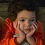 bed, bedding, blanket, casual_clothing, child, close_up, comfortable, cozy, curly_hair, face, hands_on_face, indoor, orange_shirt, pensive, person, portrait, relaxed, resting, thinking, young_person