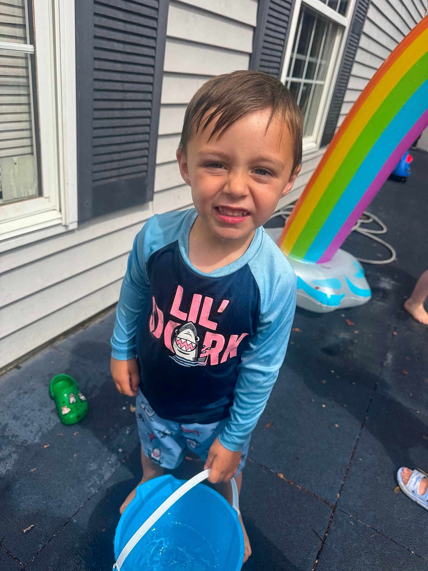 Jackson is registered to the contest to win money with this photo: child, boy, bucket, water, swimwear, rainbow, inflatable, outdoor, patio, wet_floor, window, house_siding, summer, play, barefoot, expression, fun, plastic_shoe, sunlight, daytime