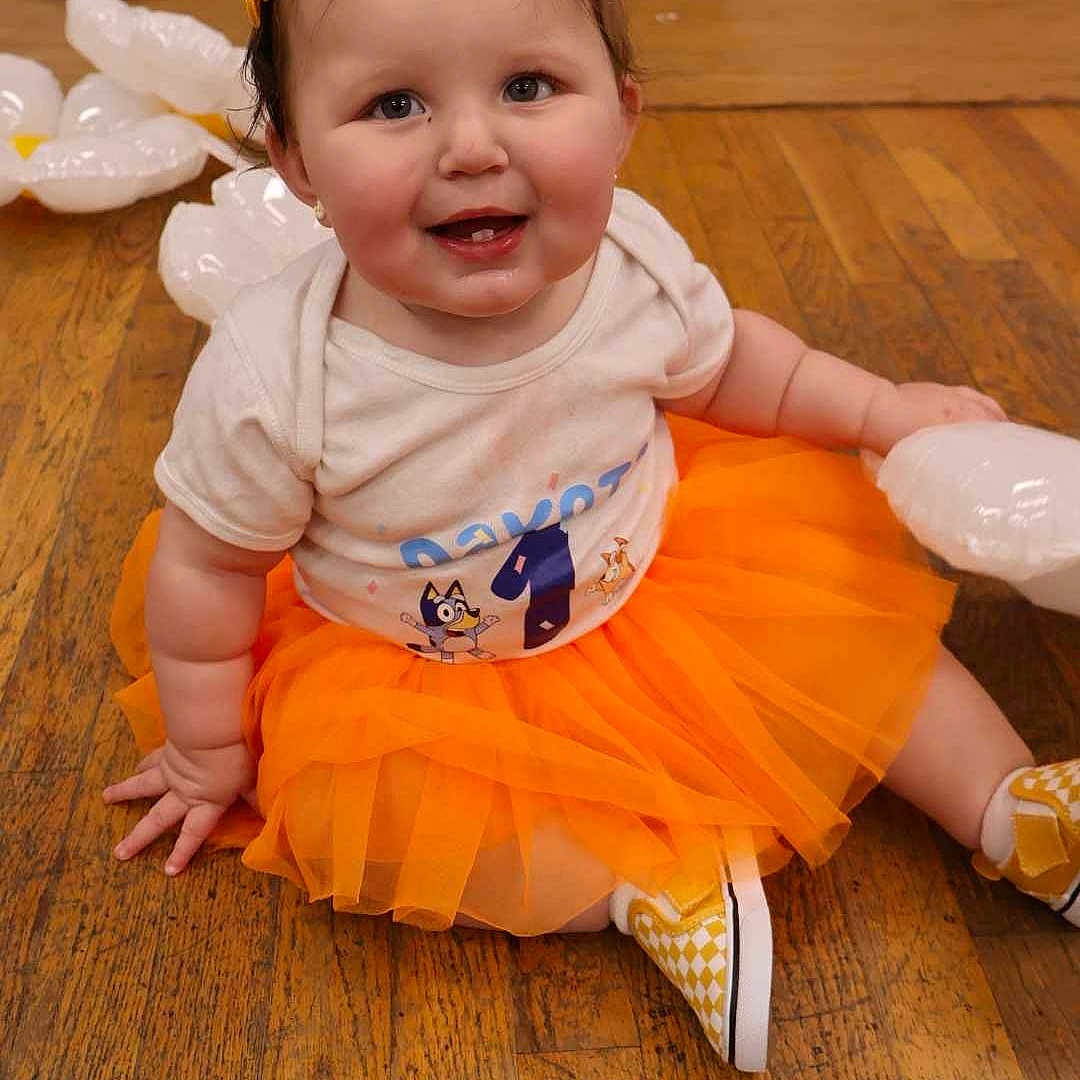 Dakota is registered to the contest to win money with this photo: accessories, baby, clothing, dress, face, footwear, happy, hardwood, head, indoors, interiordesign, jewelry, necklace, person, photography, portrait, shoe, sitting, skirt, wood