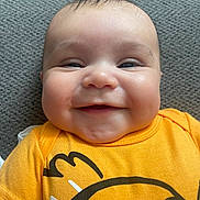 Calvin is registered to the contest to win money with this photo: baby, smiling, yellow_shirt, cartoon_print, chubby_cheeks, face, infant, cute, happy, portrait, closeup, lying_down, textile_background, short_hair, indoors, child, toddler, skin, expression, person