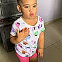 toddler, child, indoor, standing, pouty_face, hair_buns, colorful_shirt, pink_shorts, black_shoes, floor, tile_floor, person, cute, expression, hand_gesture, young_child, casual_clothing, earrings, light_wall, cabinet