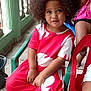 child, girl, curly_hair, red_dress, plastic_chair, indoor, railing, tiled_floor, person, sitting, portrait, casual_clothing, curious_expression, afro_hair, young_child, smiling, relaxed, home, daylight, people
