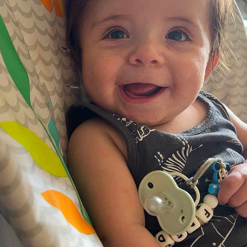 Freddie-James is registered to the contest to win money with this photo: accessory, baby, baby_seat, child, closeup, clothing, cute, face, hair, hand, happy, indoor, infant, mickey_mouse, pacifier, pattern, person, seatbelt, smiling, toy