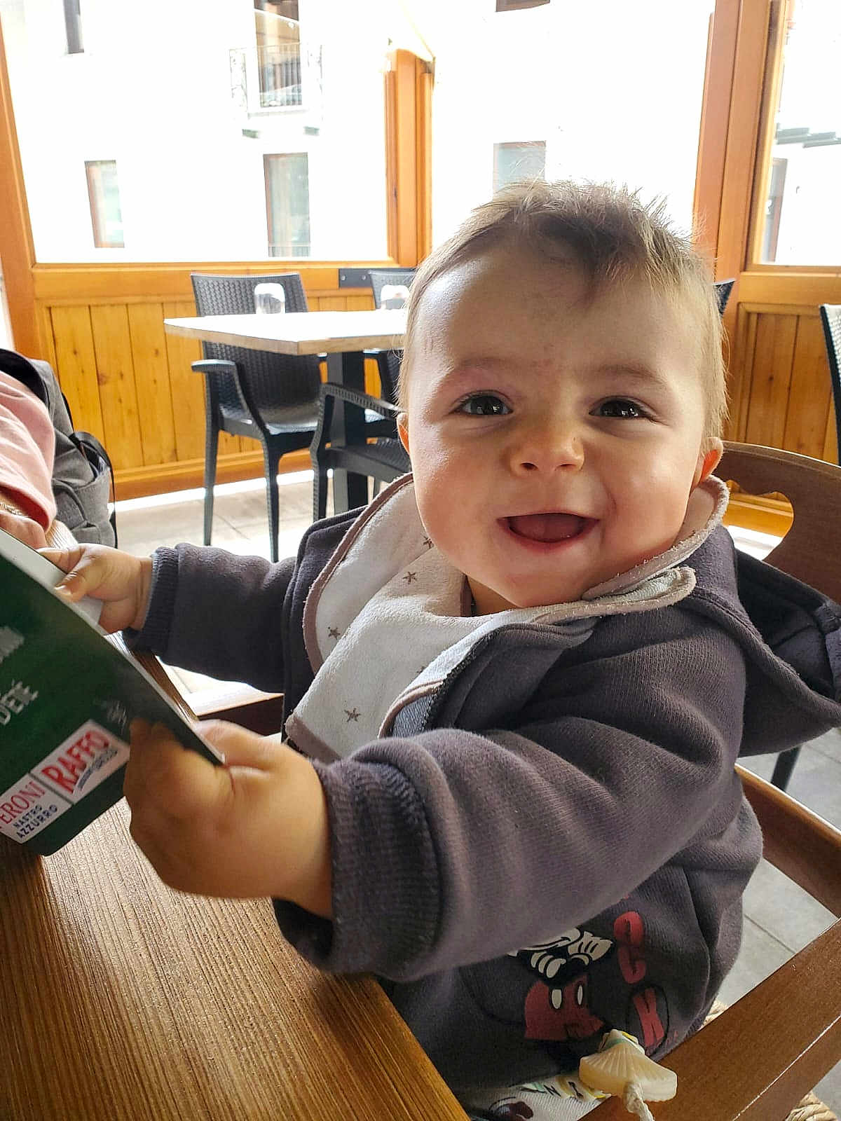 Arthur a rejoint le concours — aidez-le/la à gagner de superbes lots ! baby, child, smiling, happy, high_chair, jacket, bib, table, chair, indoor, menu, wooden_table, window, daylight, person, face, cute, infant, holding, restaurant