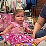 Stevie is registered to the contest to win money with this photo: baby, child, party_hat, pink_dress, toy, gift_bag, floor, indoor, person, carpet, adult_hand, celebration, party, pom_pom, face, sitting, curious, decorations, colorful, young_child