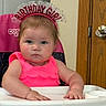 Stevie is registered to the contest to win money with this photo: baby, child, birthday_girl, high_chair, pink_dress, headband, indoor, person, face, expressionless, hands, furniture, door, wood, celebration, young_child, sitting, portrait, clothing, cute