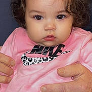 Sophia is registered to the contest to win money with this photo: adult_hands, baby, brown_eyes, child, close_up, clothing, curly_hair, cute, denim_jeans, expression, indoor, looking_at_camera, nike_logo, person, pink_shirt, portrait, sitting, skin, supporting_hands, toddler