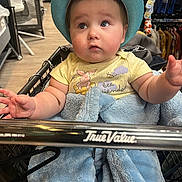 Liam is registered to the contest to win money with this photo: baby, infant, shopping_cart, hat, blanket, blue_blanket, teal_hat, chubby_cheeks, curious_expression, portrait, close_up, indoor, store, retail, clothing_rack, hands, eyes, mouth, cute, person