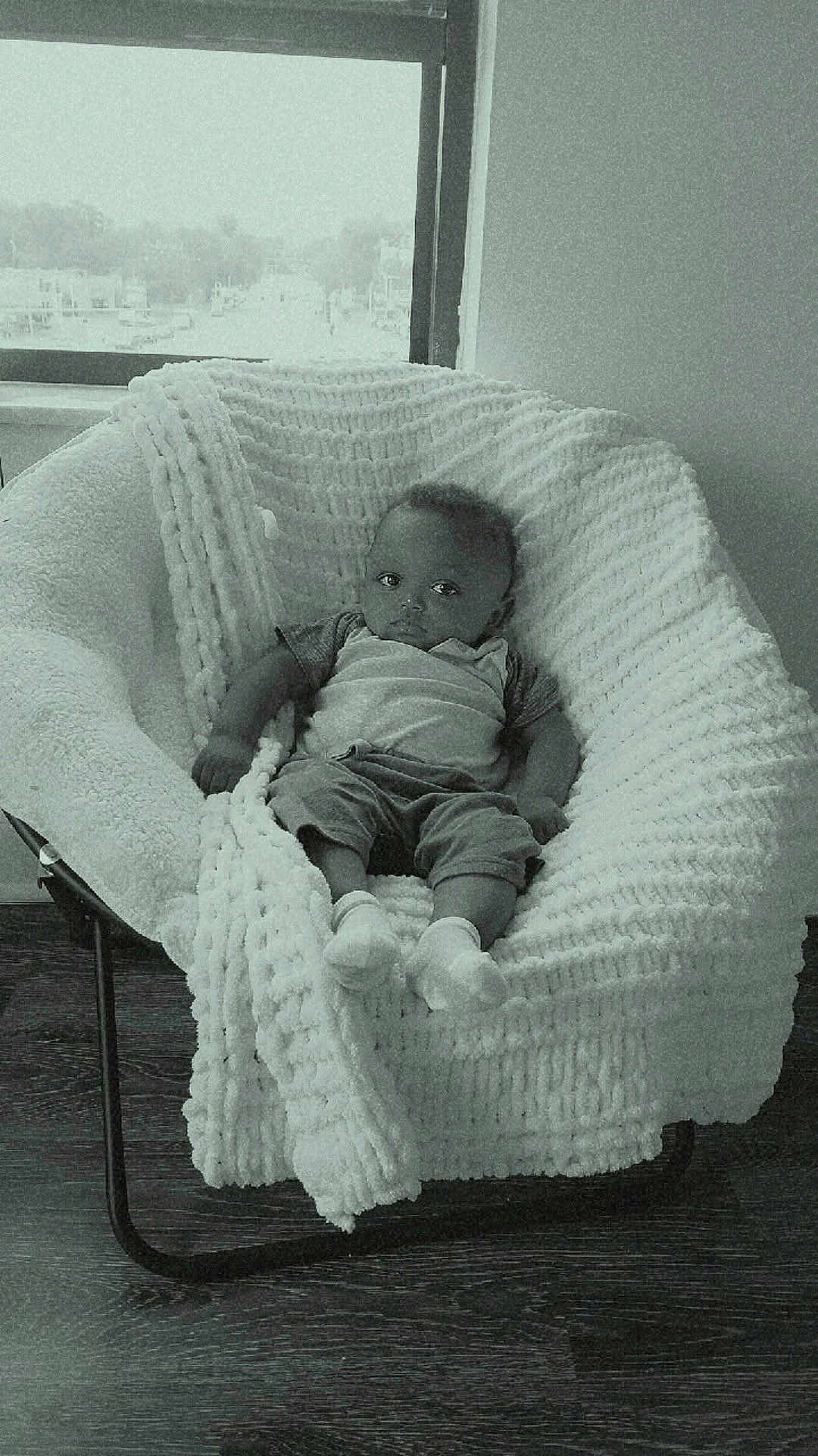 Jeremiah is registered to the contest to win money with this photo: baby, infant, blanket, chair, knitted, cozy, indoor, window, floor, wooden_floor, person, child, relaxing, casual_clothing, socks, portrait, seated, soft_texture, monochrome, calm