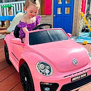 Aurora is registered to the contest to win money with this photo: child, toddler, toy_car, pink_car, outdoor, playground, playhouse, smiling, happy, summer, daylight, backyard, slide, vehicle, person, short_hair, purple_clothing, wooden_floor, sunlight, cute