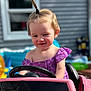 toddler, child, toy_car, purple_dress, smiling, outdoor, playtime, hair_ponytail, baby, sunlight, window, plastic_toy, happy, young_child, summer, backyard, person, steering_wheel, cute, fun
