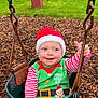 Atlas is registered to the contest to win money with this photo: baby, child, swing, playground, christmas, santa_hat, elf_costume, green, red, smiling, thumbs_up, wood_chips, outdoor, holiday, festive, happy, person, seat, fence, tree