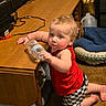baby, blanket, cables, carpet, checkered_shorts, child, curious, floor, furniture, household_items, indoor, kneeling, person, pet_bed, plastic_container, red_tank_top, room, toddler, water_bottle, wooden_table