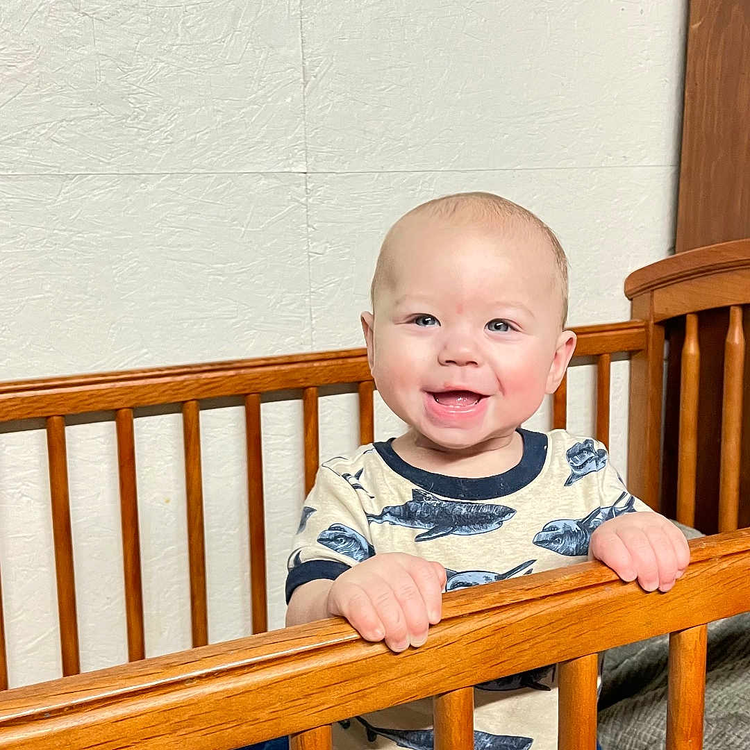 Carl is registered to the contest to win money with this photo: baby, crib, wooden_bars, smiling, child, indoor, clothing, patterned_shirt, pants, happy, face, person, furniture, bed, blanket, home, light, cute, standing, infant