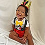 baby, smiling, crown, birthday, one_year_old, yellow_bow, red_shorts, white_shirt, sitting, happy, cute, child, black_hair, indoors, fabric_backdrop, celebration, polka_dot_suspender, portrait, infant, festive