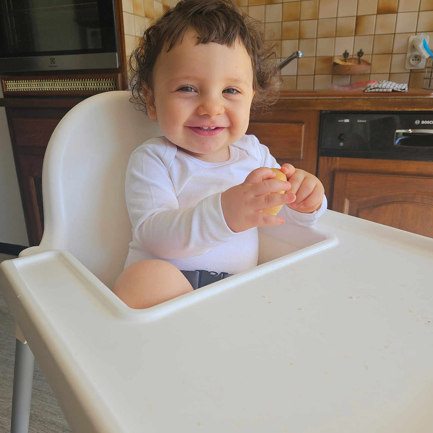 Rael a rejoint le concours — aidez-le/la à gagner de superbes lots ! baby, child, curly_hair, face, food, furniture, happy, high_chair, home, indoor, kitchen, person, seat, smiling, snack, table, tile_wall, toddler, white_clothing, wooden_cabinets